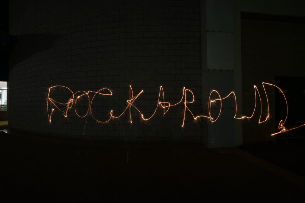 Long-exposure photo of sparkler light-painting spelling "rock and roll" in gestural orange light against a dark brick wall at night.